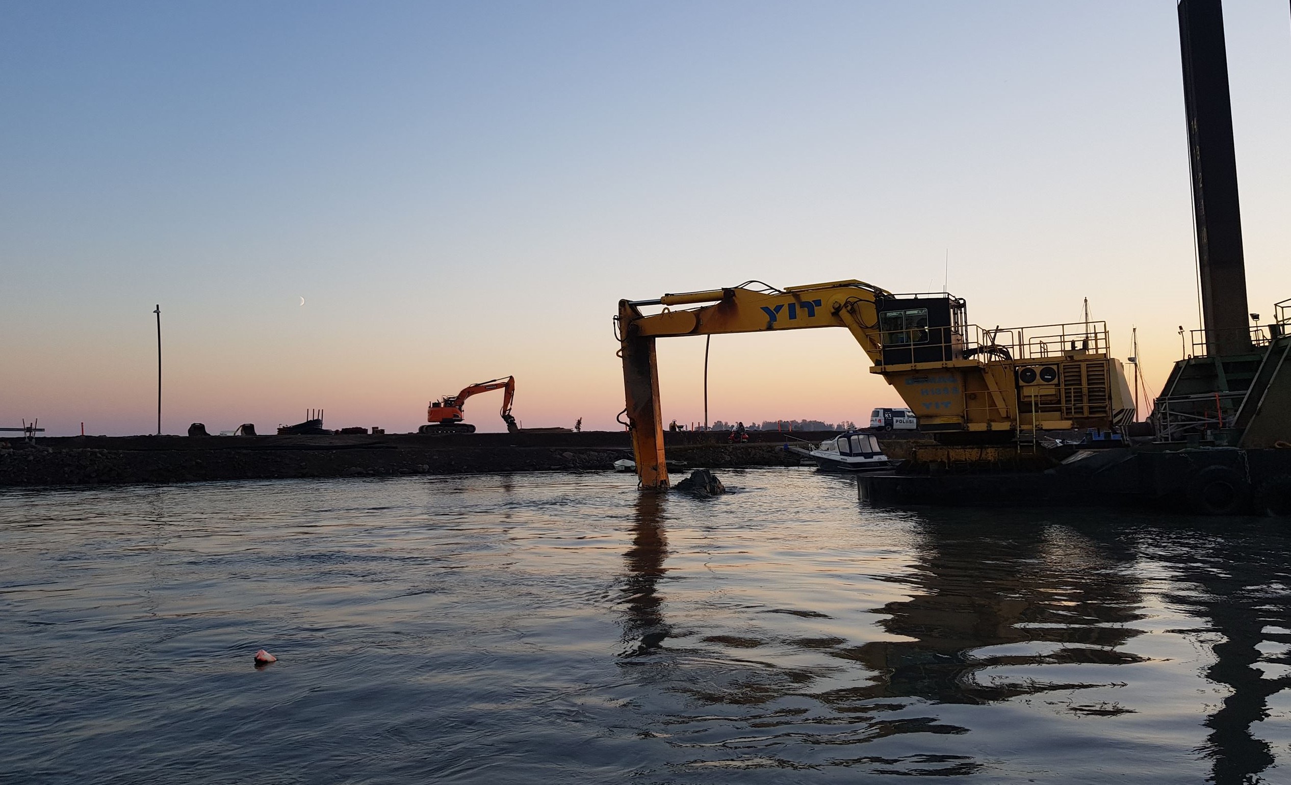 An excavator on a floating platform works in the water at sunset, with another excavator on the shoreline in the background.