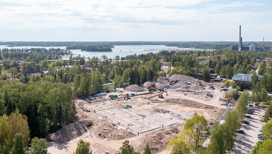 Ilmakuva Tiistilän rakennustyömaasta, jossa kaivurit tekevät maansiirtotöitä hiekkakentällä meren ja voimalaitoksen näkyessä horisontissa.